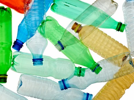An arrangement of multiple discarded plastic bottles in various colors including green, yellow, white, and blue, piled together in a haphazard manner with some bottles overlapping and leaning on each other. The bottles are made of translucent plastic, with visible caps in contrasting colors such as blue and green. The textures range from smooth to ridged, particularly on the bottle bodies, with some showing slight dents and creases typical of used containers. They are resting on an unspecified surface, which appears to be outdoors or in a warehouse environment, with no other objects or background elements visible. The lighting is even, highlighting the transparency and colors of the bottles, emphasizing the presence of recyclable waste that commercial waste removal services like Commercial Waste Osterley often handle in urban areas near Osterley or wider London suburbs. This detailed description supports accessibility needs by clearly conveying the general appearance and material of the waste material prepared for collection or disposal.