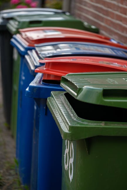 A large pile of mixed waste, including cardboard boxes, plastic bags, paper, and discarded packaging, is overflowing from multiple closed and open bins on a paved urban street. The garbage bags are bl